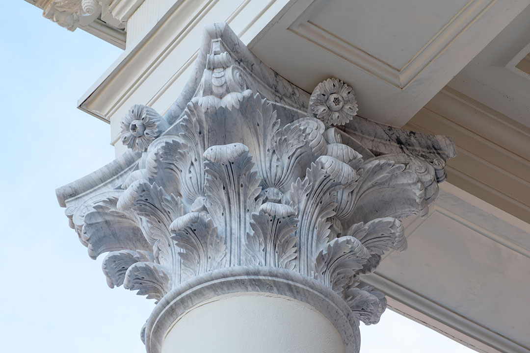 Architectural detail of a restored building facade featuring carved marble dentil molding, ornate capitals, and a contrasting red brick wall.