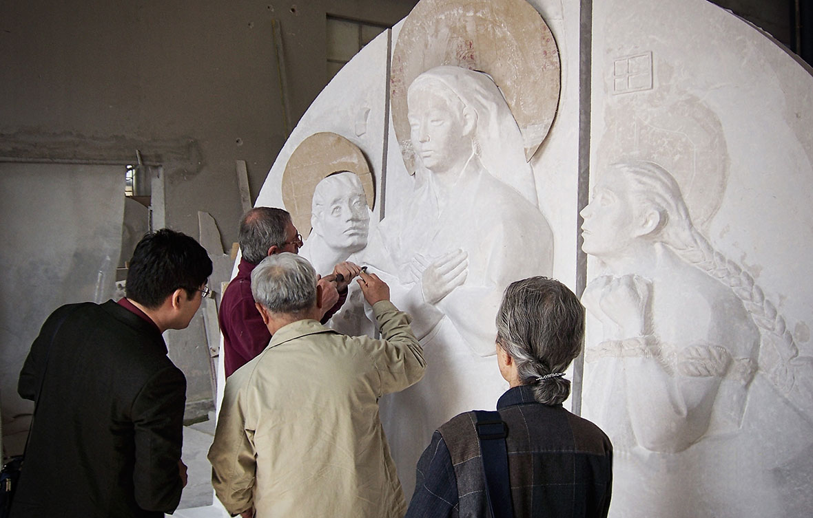 A stonemason using a power tool to refine a large-scale marble relief featuring figures in traditional Korean attire, demonstrating a blend of modern fabrication and artisan finishing.